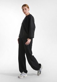 Zwarte oversized sweatshirt en bijpassende broek, gemaakt van zachte stof. Het model draagt lichtgrijze sportschoenen met paarse accenten.