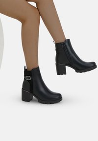 Bottes de cheville noires à talons avec une finition en cuir lisse, des côtés texturés, une fermeture éclair sur le côté et un détail de boucle à l'arrière.