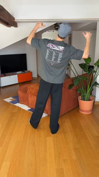 Šedé tričko s potiskem na zádech, v kombinaci s černými kalhotami. Stojí v světlé místnosti s dřevěnou podlahou a pokojovými rostlinami.