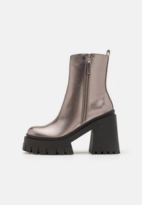 Botín plateado metálico hasta el tobillo con una plataforma y un tacón negro grueso, que presenta un zipper lateral y una textura suave y reflectante.