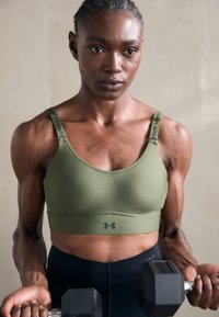 Sutien desportivo verde-oliva com alças largas, apresentando detalhes em logótipos escuros e um design sem costuras. Modelo segura um haltere preto.