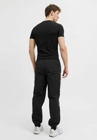 Schwarzes kurzärmliges Hemd kombiniert mit schwarzen Jogginghosen in schmaler Passform und weißen Seitenstreifen, getragen mit hellen Sportschuhen.
