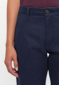 Main droite reposant à côté de la taille d’un pantalon bleu marine avec bouton et passants, portant un haut bordeaux foncé.