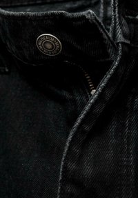 Jeans de mezclilla negra con un botón metálico y cremallera visible. La tela tiene una textura suave con un ligero brillo, con costuras en contraste.