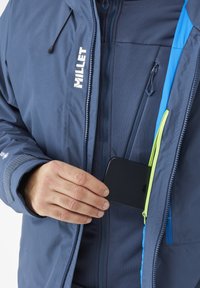 Hand sliding a black smartphone into the inner chest pocket of a blue Millet jacket with yellow zipper lining.