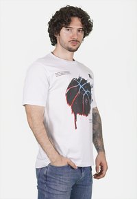 T-shirt bianca a maniche corte con una graffica di un pallone da basket che gocciola in nero, rosso e blu, con testo stampato sopra e sotto il design.
