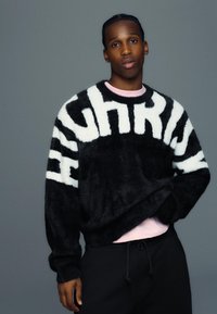 Pull noir et blanc en matière duveteuse avec le texte audacieux "HIGH" en travers du devant, porté sur une chemise rose pâle, associé à un pantalon noir.