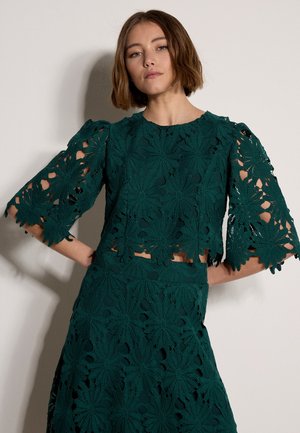 Frau trägt dunkelgrünes, florales Spitzenoberteil mit ellbogenlangen Ärmeln und passendem Rock, steht mit den Händen in die Hüften gestützt vor einer schlichten Wand.