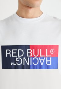 Balta T-kreklā ir drosmīgs grafiskais attēlojums ar tekstu "RED BULL RACING" melnā un sarkanā krāsā uz krāsainas fona bloka.