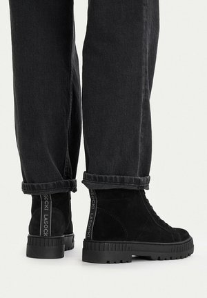 Black suede ankle boots with thick rubber soles worn with black rolled-up jeans, showing back pull tabs with brand text.