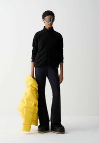 Zwarte fleecejas, gele gewatteerde jas en zwarte uitlopende broek met gestructureerde details. Draagt oversized zonnebril en dikke plateauzolen.