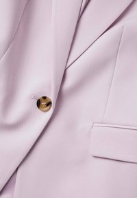Gros plan sur un blazer violet clair montrant le revers, un bouton en écaille de tortue et le détail de la poche à rabat sur un tissu lisse.