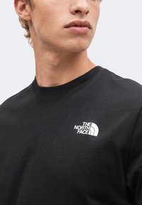 Νέος/νεαρό άτομο που φοράει μαύρο t-shirt με στρογγυλή λαιμόκοψη και λευκό λογότυπο "The North Face" στην αριστερή πλευρά του στήθους, κοιτάζοντας προς τα πλάγια.