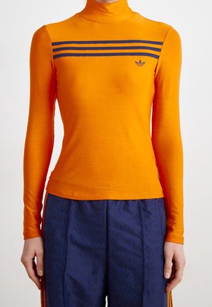 Personne portant un col roulé orange à manches longues avec trois bandes bleu marine et un logo sur la poitrine, associé à un pantalon bleu marine à motifs.