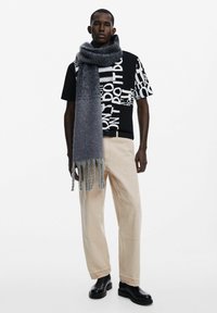 Camiseta gráfica negra con texto blanco, pantalones beige de pierna ancha, bufanda de punto gris con flecos y botines negros.