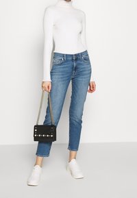 Femme portant un pull col roulé blanc à côtes, un jean bleu cropped, des baskets blanches, tenant un sac à main noir orné de perles et avec une bandoulière en chaîne.