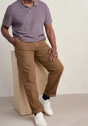 Mann mit einem lila kurzärmeligen Poloshirt, beigen Hosen, schwarzen Socken und weißen Sneakers sitzt auf einem Holzblock mit einer Hand in der Tasche.