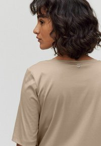 Beige t-shirt med kort ärm som har en rund halsringning och en liten metallakcent bak. Mjuk, slät tygkvalitet med en avslappnad passform.