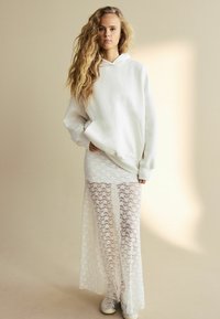 Bílá oversized mikina s klokankou, v kombinaci s dlouhou bílou krajkovou sukní se květinovými vzory a rozparky, doplněná stříbrnými teniskami.