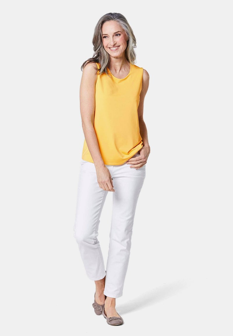 Top giallo senza maniche, tessuto liscio, scollatura rotonda, abbinato a pantaloni in denim bianchi e scarpe piatte grigie con dettaglio a fiocco.