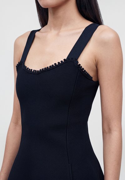 Gros plan d'une femme portant une robe noire sans manches avec de larges bretelles et une bordure décorative le long de l'encolure.