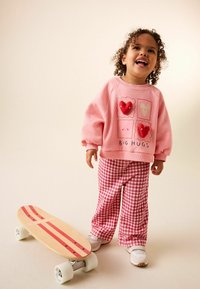 Felpa rosa con dettagli a forma di cuore e scritta "BIG HUGS"; pantaloni a quadretti rosso e bianco; skateboard di legno con strisce rosse.