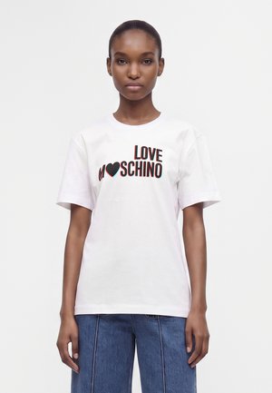 Γυναίκα που φοράει λευκό μπλουζάκι Love Moschino με μαύρο και κόκκινο κείμενο και μπλε τζιν, στέκεται μπροστά σε απλό λευκό φόντο.