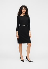 Robe noire longueur genou avec manches trois-quarts, encolure en V et une fine ceinture noire. Portée avec des talons noirs. Texture lisse.