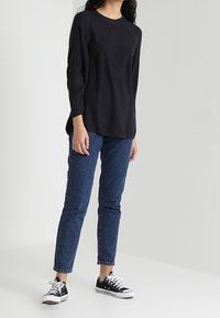Schwarzes langärmeliges Rippoberteil kombiniert mit blauen geraden Jeans und schwarzen Converse-Sneakern vor hellen Hintergrund.