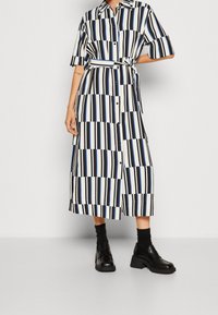 Abito camicia con bottoni, maniche corte, a righe verticali bianche e nere con accenti blu. Presenta un cintura aderente in vita e lunghezza fino al polpaccio.