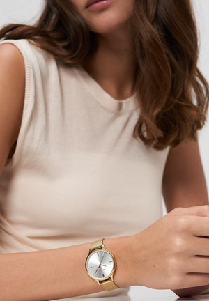 Jeune femme portant un haut beige sans manches avec une montre-bracelet à bracelet en maille dorée et un cadran argenté à son poignet gauche.