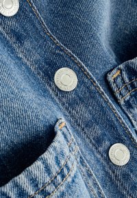 Lähikuva sinisestä denimpainokankaan kankaasta, jossa on oranssia ommelta ja hopeanvärisiä nappikiinnityksiä, merkitty "Next Essential Jeanswear."