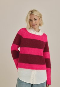 Pletený sveter s širokými horizontálnymi pásmi v fuksiovej a tmavej burgundskej farbe, s jemnou textúrou, položený na bielu košeľu s golierom.
