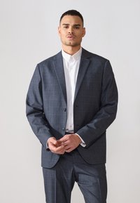 Abito a scacchi blu navy con rever a punta, giacca monopetto e pantaloni coordinati. Camicia bianca con bottoni indossata sotto.