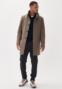 Brauner Wollmischungs-Mantel mit hohem Kragen, schwarzen Knöpfen und seitlichen Taschen, kombiniert mit schwarzen Hosen und beigen Sportschuhen.