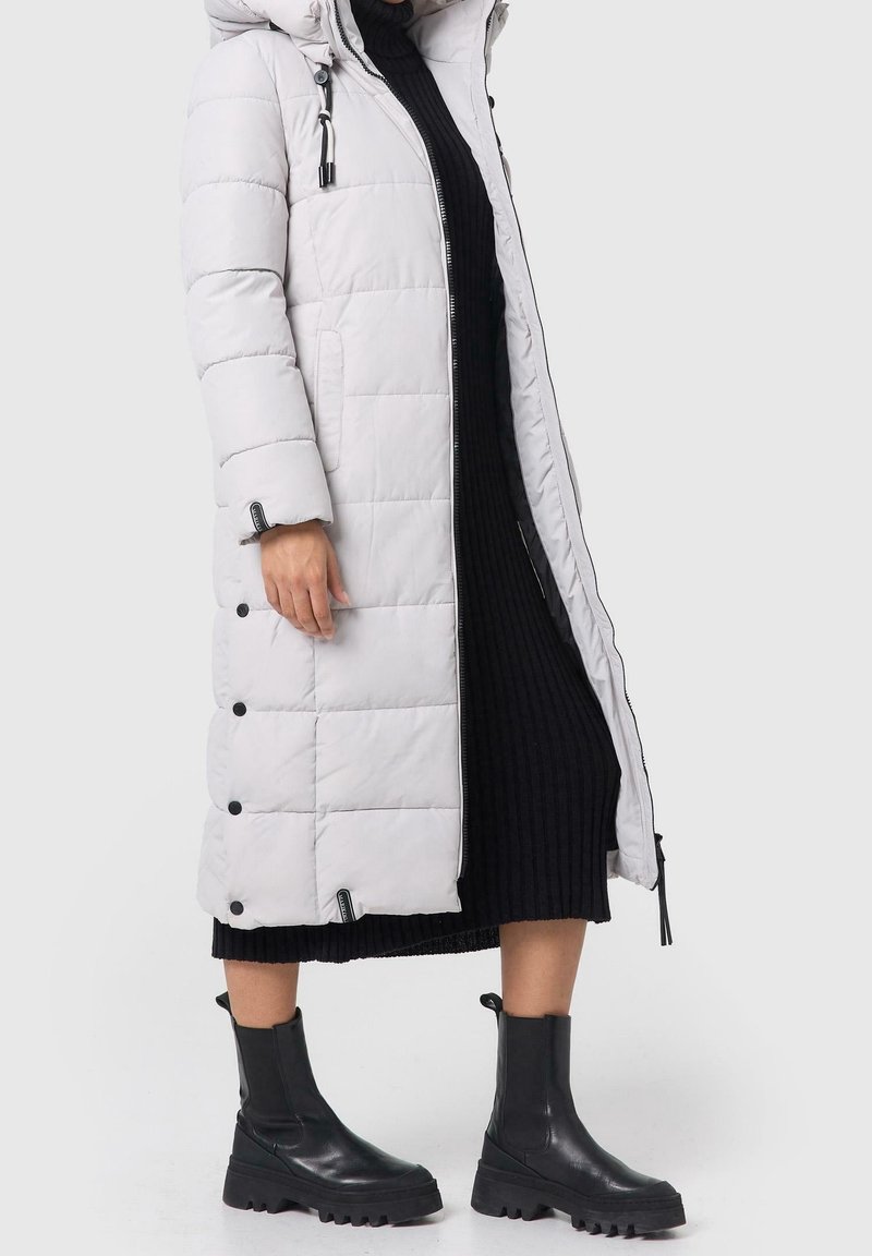 Femme portant un long manteau matelassé gris clair sur une robe noire côtelée et des bottines noires épaisses, debout devant un fond uni.