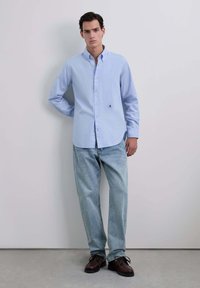 Chemise à manches longues bleu clair avec un petit logo brodé, associée à un jean bleu délavé et des chaussures marron, se tenant contre un mur neutre.