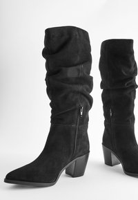 Bottes noires en daim jusqu'aux genoux avec un design froissé, un bout pointu et un talon carré. Équipées de fermetures éclair latérales pour un enfilage facile.