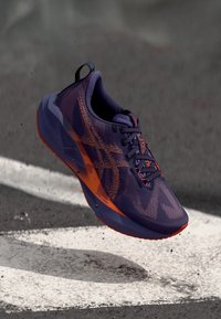 Zapatilla para correr morada con detalles en naranja, parte superior texturizada, diseño aerodinámico y suela acolchada. Presenta un logo en el lateral y cordones.