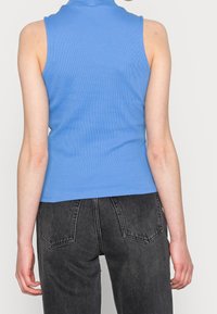 Ljust blå ärmlös ribbad topp med hög halsdesign, som har en figurnära passform och en mjuk textur, tillsammans med högmidjade mörka jeans.