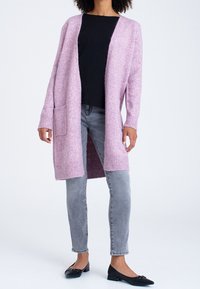 Lång, öppen cardigan i mjuk rosa stickning med ribbade ärmslut och nederkant, med en framficka. Matchas med grå smala jeans och svarta flats.