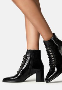 Botine negre din piele patentată, cu vârf ascuțit, finisaj texturat, fermoar lateral și detalii decorative cu șireturi în față.