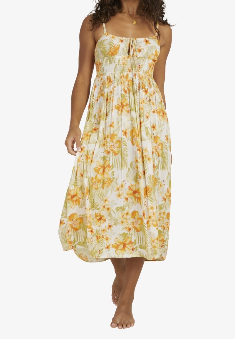 Femme marchant pieds nus, portant une robe d'été midi blanche à imprimé floral jaune et vert avec des bretelles fines.