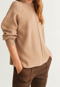 Femme portant un pull beige côtelé aux manches retroussées et un pantalon à carreaux marron, les mains dans les poches, sur un fond uni.