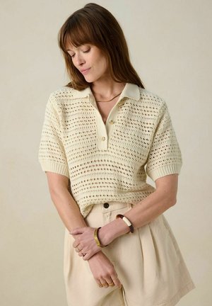 Knit polo shirt in cream with short sleeves, featuring a textured open weave, button front, and a ribbed collar. Paired with beige shorts.