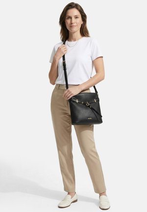 Vrouw die een wit T-shirt en beige broek draagt, houdt een zwarte leren schoudertas met gouden details vast, staand tegen een effen achtergrond.