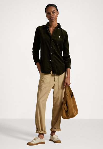 Dark green corduroy button-down shirt, beige cargo pants, cream sneakers, and a brown suede bag. Model poses against a plain background.