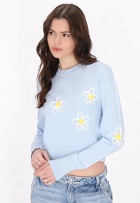 Maglione corto in blu chiaro realizzato in tessuto morbido, con applicazioni di margherite bianche e gialle sulla parte frontale e sulle maniche, dotato di polsini a costine.