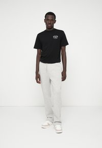 Schwarzes Baumwoll-T-Shirt mit Logo, kombiniert mit hellgrauen Sweatpants aus weichem Stoff. Das Modell trägt weiße und beigefarbene Sneakers.