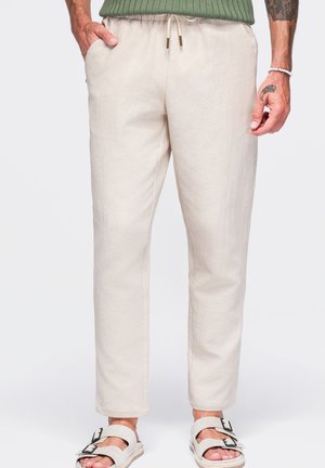 Hombre con pantalones texturizados color blanco roto con cintura ajustable, suéter verde acanalado, sandalias blancas y tatuajes en las manos y antebrazos visibles.